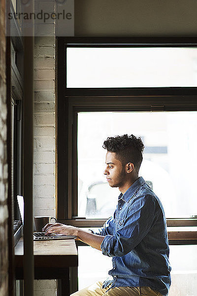 Seitenansicht eines Mannes  der seinen Laptop bei einer Kaffeetasse benutzt  während er an den Fenstern eines Cafés sitzt
