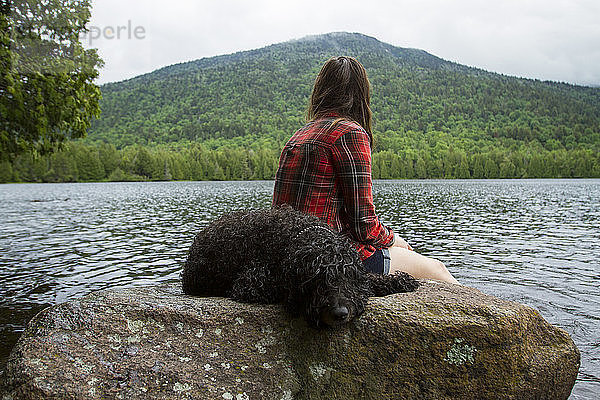 Seitenansicht einer Frau  die auf einen Teich schaut  während sie neben einem Hund sitzt  der auf einem Felsen im Wald schläft