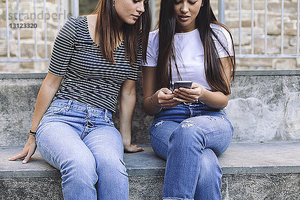 Freundinnen  die ein Mobiltelefon benutzen  während sie auf Stufen an der Wand sitzen