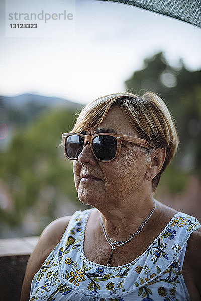 Nahaufnahme einer älteren Frau mit Sonnenbrille  die auf einer Gebäudeterrasse gegen den Himmel steht