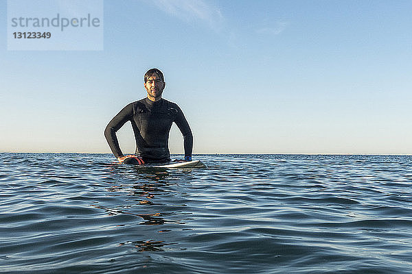 Männlicher Surfer sitzt auf einem Surfbrett im Meer vor klarem Himmel