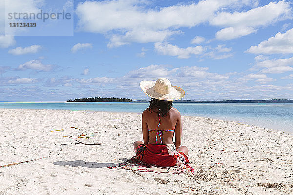 Rückansicht einer Frau mit Sonnenhut  die am Strand gegen den Himmel sitzt