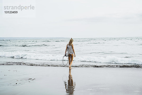 Rückansicht einer Frau  die ein Surfbrett trägt  während sie in Richtung Meer gegen den Himmel läuft