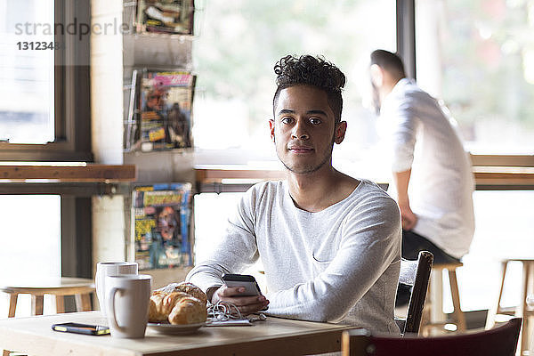 Porträt eines jungen Mannes mit Handy in der Hand bei Croissant und Kaffee im Cafe