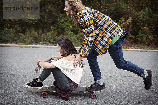 Mann schiebt Frau auf Skateboard sitzend auf Straße