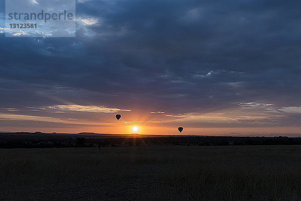 Silhouette von Heißluftballons  die bei Sonnenuntergang im Serengeti-Nationalpark vor stürmischen Wolken über die Landschaft fliegen