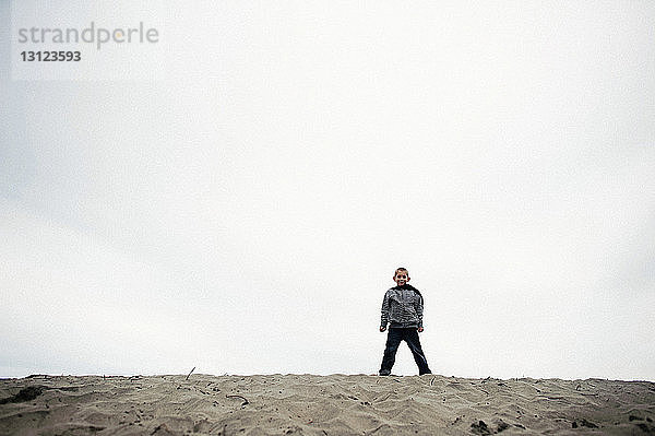 Porträt eines lächelnden Jungen  der auf Sand am Strand gegen den Himmel steht