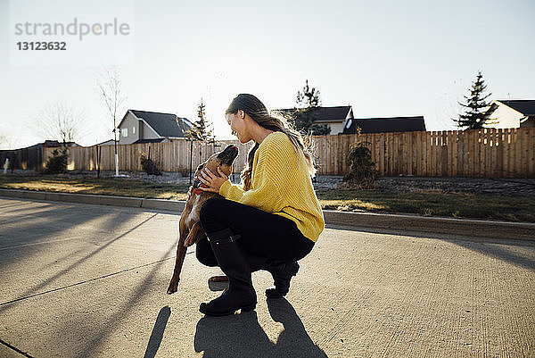 Seitenansicht einer Frau  die mit einem Hund spielt  während sie auf der Straße vor dem klaren Himmel kauert
