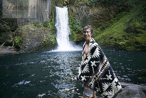 Porträt eines glücklichen Mannes im Tuch  der am Wasserfall im Wald steht