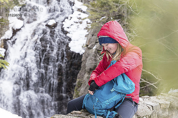 Lächelnde Frau mit Rucksack sitzt im Winter am Wasserfall in den White Mountains