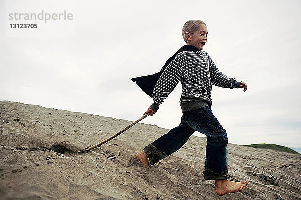 Verspielter Junge mit Stock  der auf Sand am Strand gegen den Himmel läuft