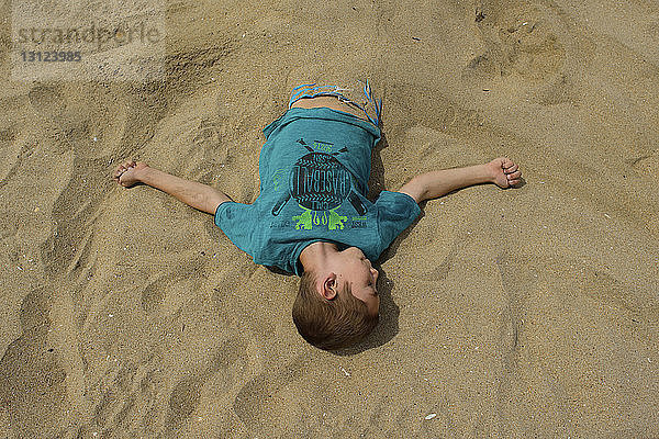 Schrägaufnahme eines am Strand im Sand begrabenen Jungen