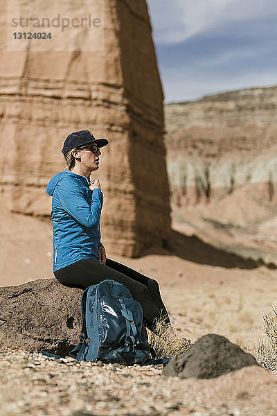 Seitenansicht einer Wanderin mit Rucksack  die sich bei Sonnenschein auf einem Felsen in der Wüste entspannt