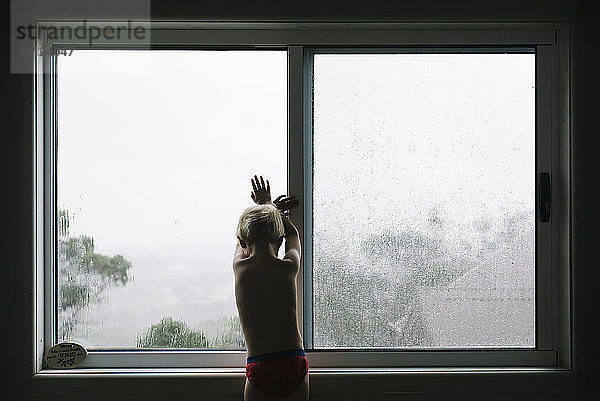 Rückansicht eines hemdlosen Jungen  der zu Hause am Fenster steht