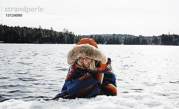 Fröhliche Frau trägt Pelzmantel  während sie im Winter im Algonquin-Provinzpark in eine Decke gegen den See gewickelt wird