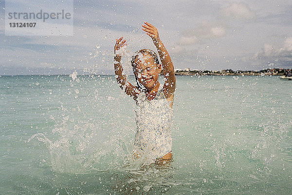 Fröhliches Mädchen plätschert Wasser im Meer gegen bewölkten Himmel