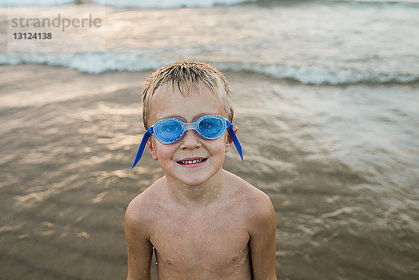 Porträt eines Jungen ohne Hemd mit Schwimmbrille  der am Strand am Ufer steht