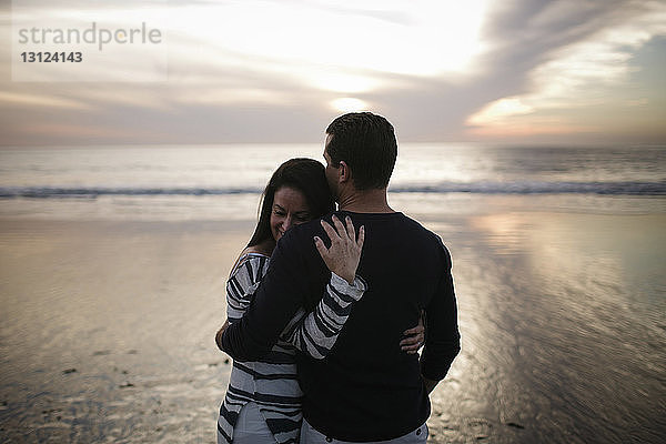 Paar umarmt sich  während es bei Sonnenuntergang am Strand vor bewölktem Himmel steht