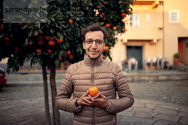 Porträt eines Mannes  der eine Orange hält  während er an einem Baum auf einem Fußweg steht