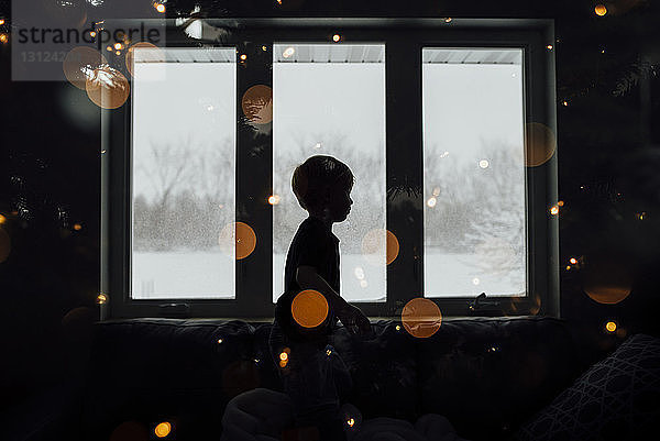 Seitenansicht eines am Fenster stehenden Silhouettenjungen in einem beleuchteten Raum zu Hause