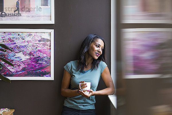 Nachdenkliche Frau hält Kaffeetasse in der Hand  während sie zu Hause an Bilderrahmen an der Wand steht