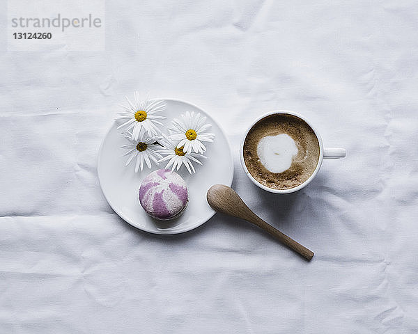 Draufsicht auf Makrone mit Gänseblümchen im Teller von Kaffee auf weißem Textil