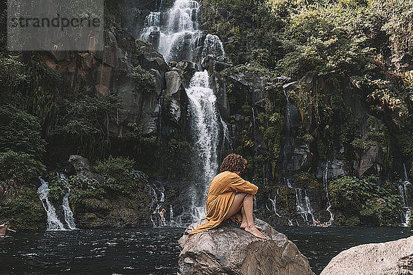 Seitenansicht einer Frau  die auf einem Felsen gegen einen Wasserfall im Wald sitzt