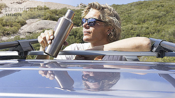 Mann mit Sonnenbrille trinkt Wasser  während er sich auf Auto stützt