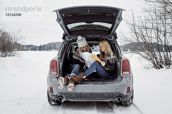 Glückliche Freundinnen  die im Winter im Kofferraum eines Autos sitzen und Karten lesen