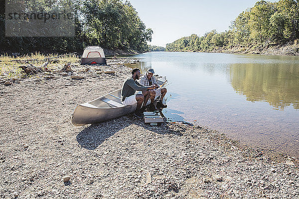 Männliche Freunde unterhalten sich beim Anpassen von Angelgeräten auf einem Boot am Seeufer