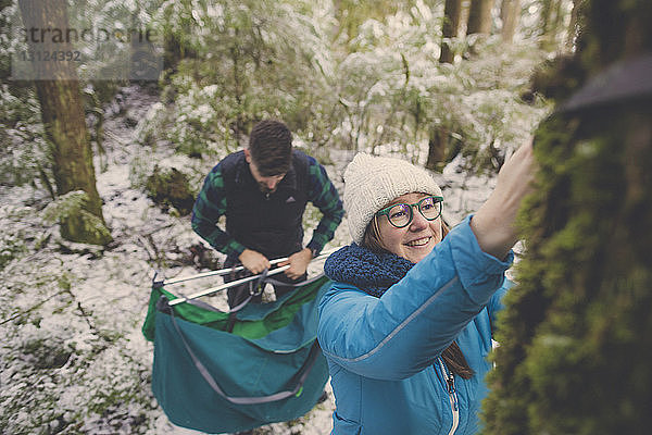 Lächelnde Frau bindet Riemen vom Baumstamm ab  während ein Mann im Wald im Lynn Canyon Park im Winter eine Hängematte hält