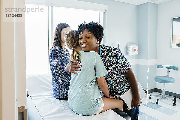 Kinderarzt umarmt Mädchen  das von der Mutter auf dem Untersuchungstisch im Krankenhaus sitzt