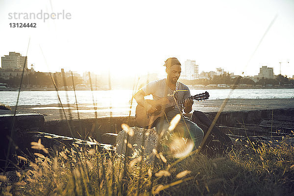 Mann spielt Gitarre  während er am Seeufer sitzt