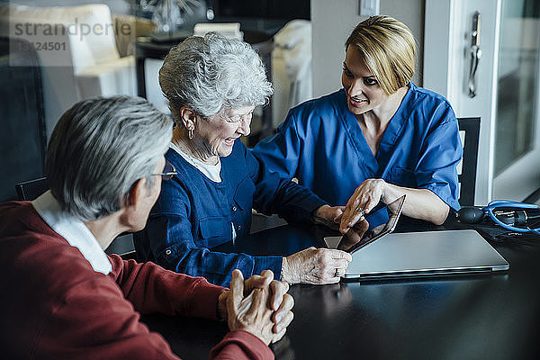 Heimpflegekraft diskutiert am Esstisch über Tablet-Computer mit älterem Ehepaar
