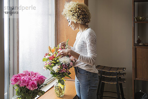 Lächelnde junge Frau arrangiert Blumen in Container auf der Fensterbank zu Hause