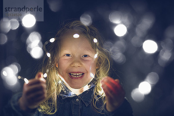 Porträt eines fröhlichen Mädchens  das nachts mit beleuchteten Lichterketten spielt