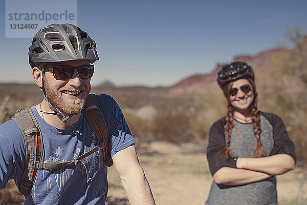 Glückliche männliche und weibliche Mountainbiker stehen am Berg vor strahlend blauem Himmel