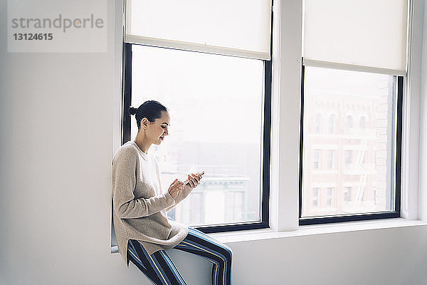 Frau benutzt Mobiltelefon  während sie zu Hause auf dem Fensterbrett sitzt