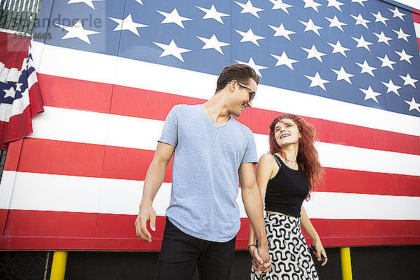 Junges Paar hält sich an der Hand  während es gegen die amerikanische Flagge steht