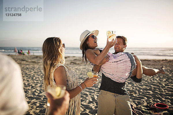 Fröhlicher Mann trägt Freundin mit Trinkglas am Strand
