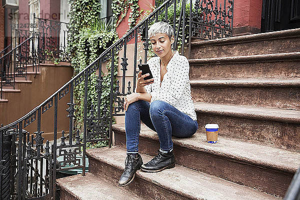 Frau benutzt Mobiltelefon  während sie mit Kaffeetasse auf der Treppe sitzt