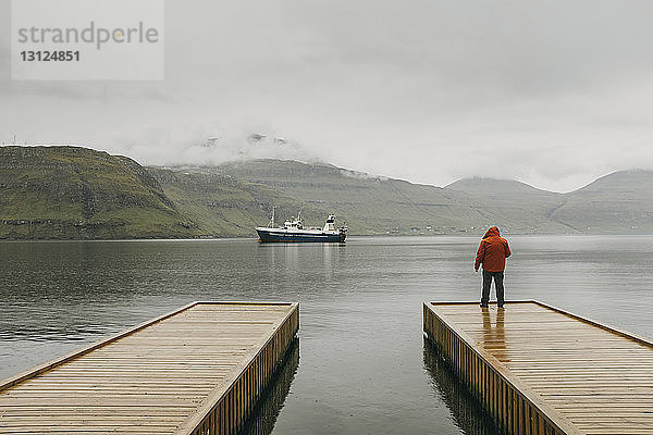 Rückansicht eines Mannes in Kapuzenjacke  der bei nebligem Wetter auf dem Pier gegen die Berge steht