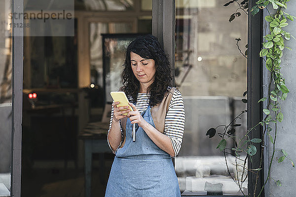 Selbstbewusste Kunsthandwerkerin benutzt Smartphone  während sie in der Werkstatt an der Tür steht