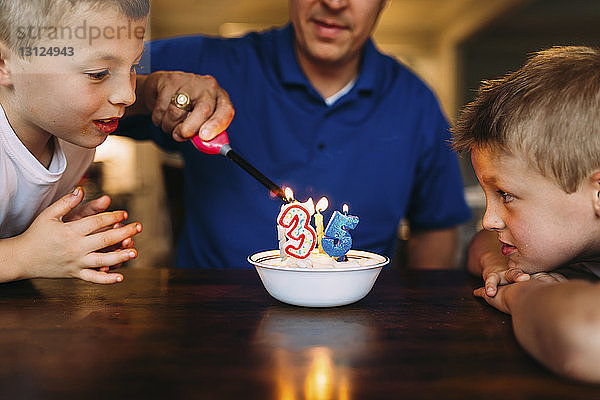 Neugierige Brüder sehen Vater beim Anzünden von Geburtstagskerzen mit Feuerzeug auf dem Tisch zu Hause