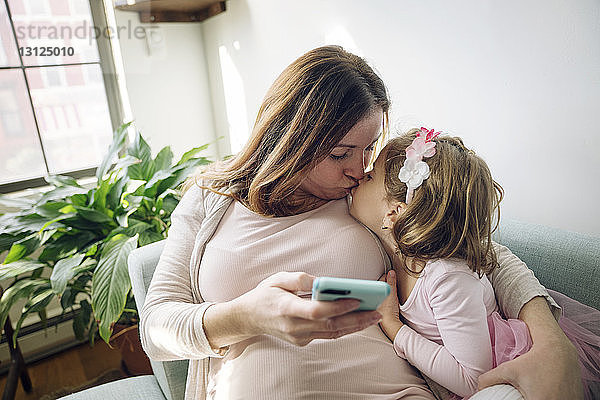 Mutter küsst Tochter  während sie zu Hause ein Smartphone in der Hand hält