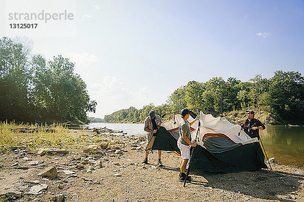 Männliche Freunde versammeln Zelt auf einem Campingplatz am See gegen den Himmel