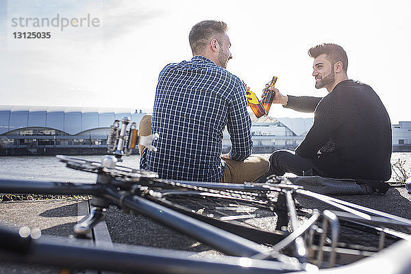 Freunde stoßen auf eine Bierflasche an  während sie mit dem Fahrrad auf der Promenade vor klarem Himmel sitzen