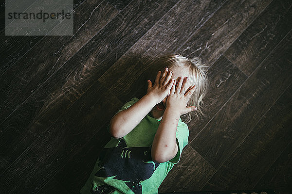 Draufsicht auf einen Jungen  dessen Hände die Augen bedecken und der zu Hause auf dem Boden liegt