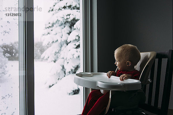 Glücklicher kleiner Junge  der im Winter zu Hause auf einem Hochstuhl sitzend durch das Fenster schaut