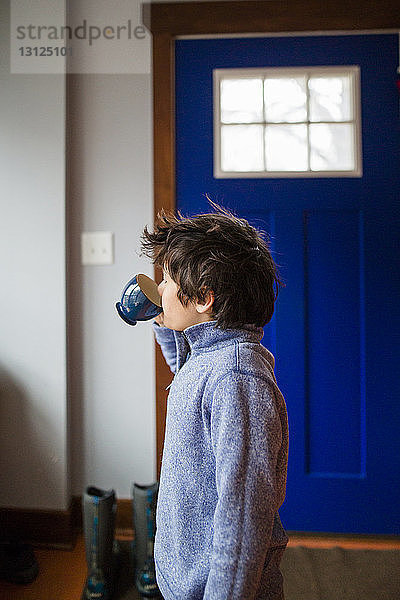 Seitenansicht eines zu Hause stehenden Jungen  der Kaffee aus einer Tasse trinkt